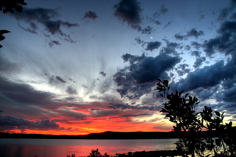 Über einem See leuchtet Abendrot, der Himmel darüber ist klar mit lebendigen Wolken, zwischen den Wolken leuchtet der Sichelmond und die Planeten Venus und Jupiter.
