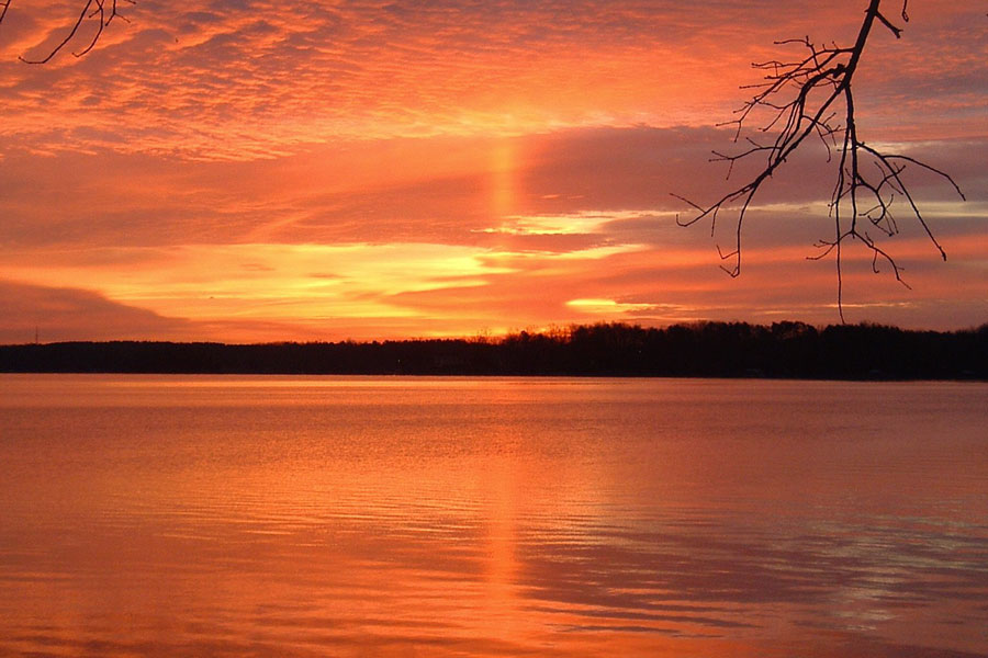 Das Bild ist leuchtend orange gefärbt, über einem See ist ein dramatischer bewölkter Himmel, am Horizont steigt eine Lichtsäule auf, die sich im Wasser spiegelt.