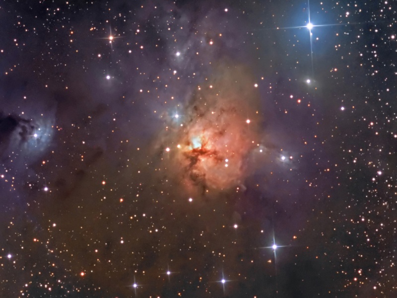 Das Sternenfeld zeigt in der Mitte einen rot leuchtenden Nebel mit Staubbahnen, darum herum sind einige kleinere blaue Nebel verteilt.
