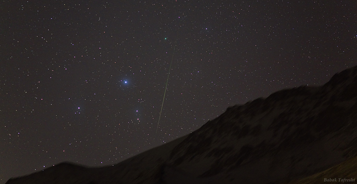 Rechts zeichnet sich am dunklen Himmel die noch dunklere Silhouette eines Berges ab, links neben der Mitte ragt das Sternbild Orion teilweise über den Horizont. Rechts daneben verläuft senkrecht eine Meteorspur.