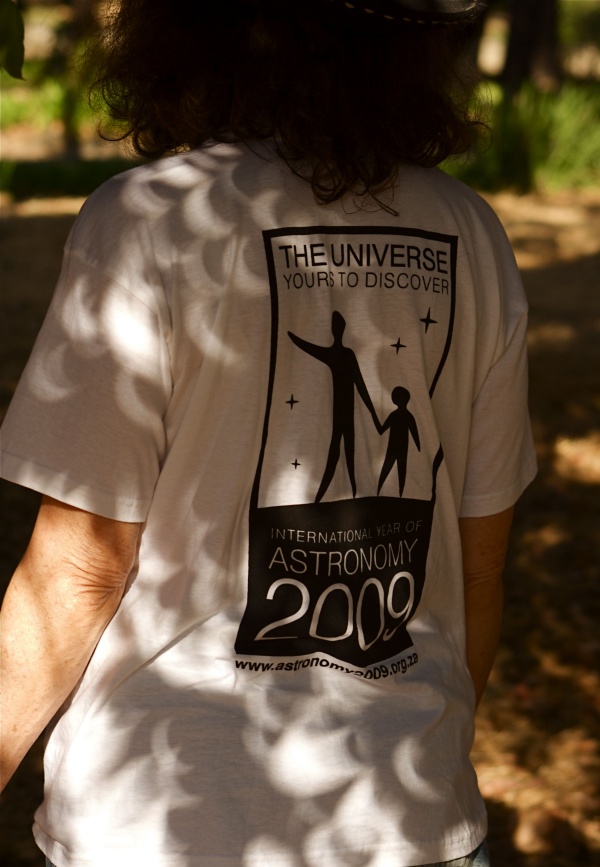In einem sonnigen Park trägt eine Frau ein weißes T-Shirt mit dem Logo des IYA 2009, darauf sind sichelförmige Sonnenbilder von einer partiellen Sonnenfinsternis.