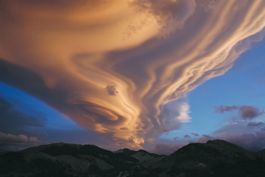 An einem wolkigen blauen Himmel verläuft quer eine orange beleuchtete geschichtete Wolkenfront. Hinten sind die Silhouetten von Bergen zu sehen.