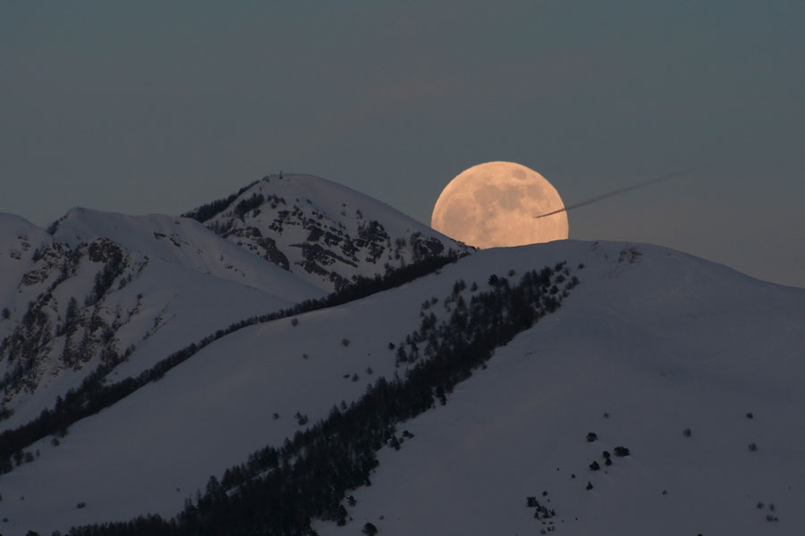 Über schneebedeckten Bergen geht der Vollmond auf. Scheinbar wird er von einem Flugzeug getroffen.