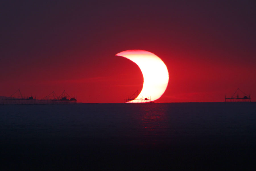 Über einem dunklen Horizont mit den Silhouetten eines Hafens strahlt eine sichelförmige Sonnenfinsternis am roten Himmel.
