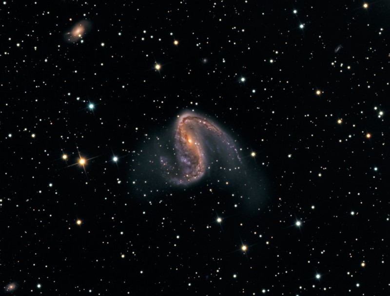Mitten im Bild mit dünn verstreuten Sternen befindet sich eine verzerrte Galaxie mit zwei markanten, asymmetrischen Spiralarmen.