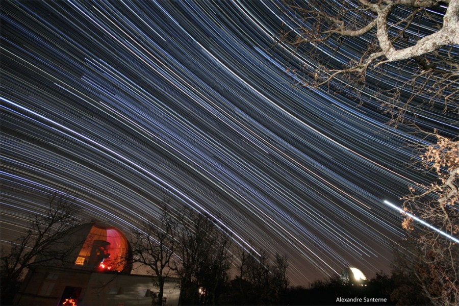 Diagonal verlaufen Strichspuren von Sternen durchs Bild, links unten ist die geöffnete, innen rot beleuchtete Kuppel einer Sternwarte zu sehen, rechts sind die Äste von Bäumen.