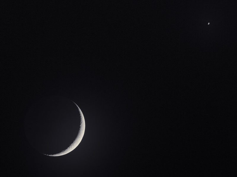 Vor einem schwarzen Hintergrund sind zwei Sicheln zu sehen: Links unten der Mond, rechts oben winzig klein die Venus.