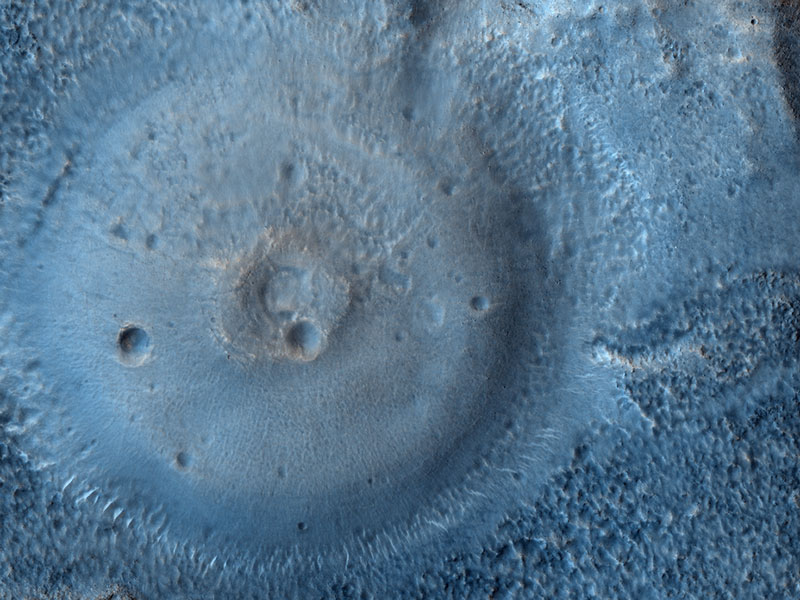 In der linken Bildhälfte ragt eine runde Struktur hoch, die in der Mitte eine Art Krater besitzt, der wiederum einen Einschlagkrater besitzt. Das Bild ist hellblau gefärbt.