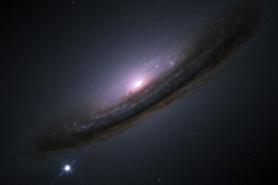 Schräg im Bild liegt eine von seitlich oben sichtbare Galaxie mit einem sehr dunklen, markanten Staubring. Das Zentrum leuchtet hell. Links unten ist ein helles Licht, eine Supernova.