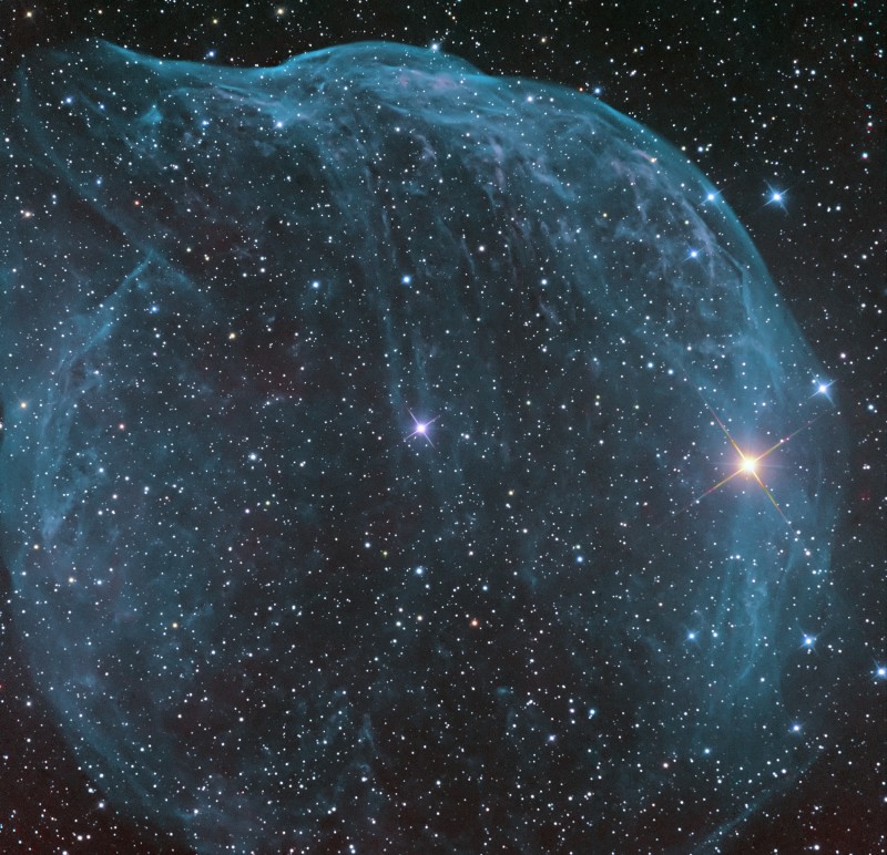 Das Bild zeigt einen blauen, blasenartigen Nebel, der fast das ganze Bild ausfüllt. Aus dem Sternenteppich leuchten in der Mitte und rechts zwei hellere Sterne hervor.