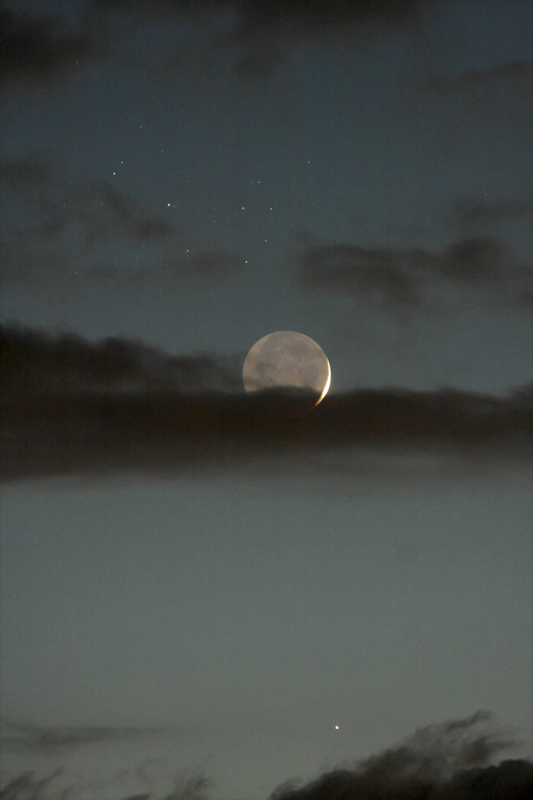 An einem dunklen taubenblauen Himmel ist der zarte Sichelmond unten von einem Wolkenstreifen bedeckt, die Nachtseite ist von der Erde beleuchtet. Unten leuchtet der Planet Merkur.
