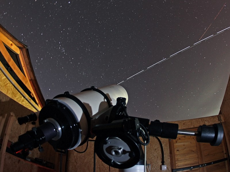 Ein Teleskop in einer geöffneten Hütte ist auf den Sternenhimmel gerichtet, über den die Internationale Raumstation zieht.