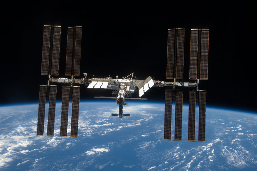 Über der blauen Erde mit Ozeanen und wenigen Wolken schwebt die Internationale Raumstation mit mächtigen Solarpaneelen.