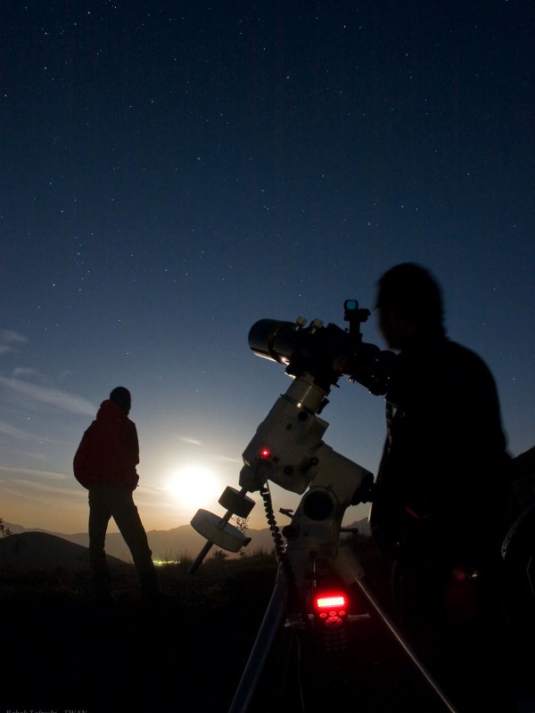 Zwei Silhouetten und ein Teleskop zeichnen sich vor einem klaren dunkelblauen Himmel ab, über dem Horizont leuchtet ein sehr helles Himmelslicht.