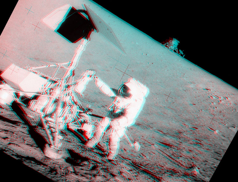 Das Bild wurde schräg fotografiert. Im Vordergrund steht ein Astronaut bei einer Raumsonde, im Hintergrund am Horizont steht das Landemodul. Mit rot-grünen Brillen wirkt das Bild dreidimensional.