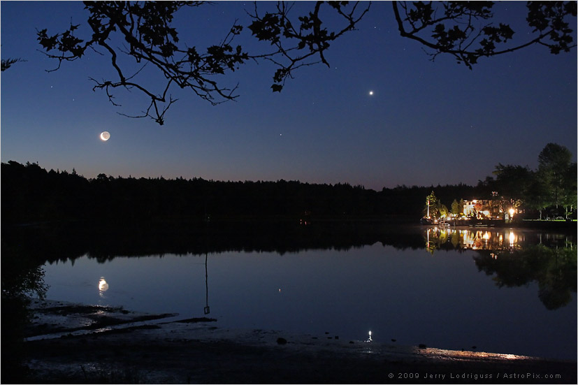 Hinter einem ruhigen Gewässer steht ein beleuchtetes Gebäude am Ufer, am dunkelblauen Himmel leuchtet ein Sichelmond mit Altlicht und der Planet Venus, beide spiegeln sich im Wasser.