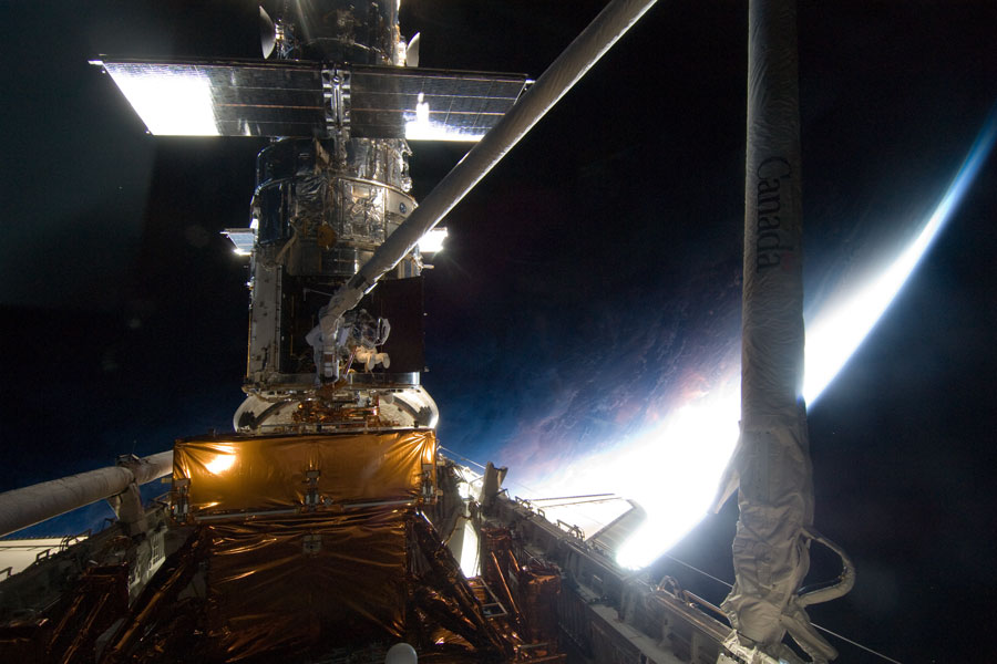 Hinter einer Ladebucht derRaumfähre Atlantis ist das Weltraumteleskop Hubble befestigt, dahinter ist eine beleuchtete Sichel der Erde zu sehen.
