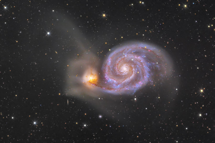 In der Bildmitte leuchtet eine Doppelspiralgalaxie, links ein kleiner, gelb leuchtender Teil, rechts eine Spiralgalaxie mit klar ausgeprägten Spiralarmen.