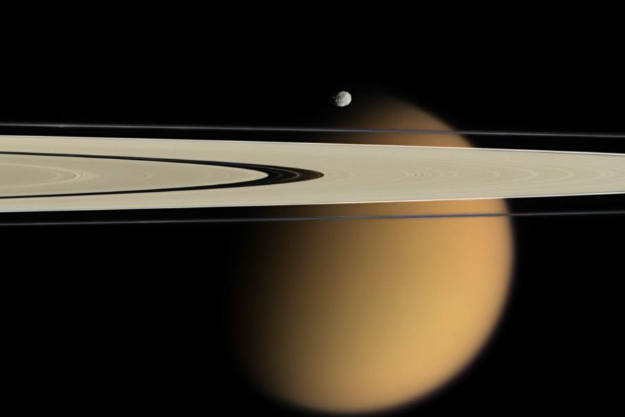 Hinter einem Asschnitt der Saturnringe mit deutlich erkennbaren Teilungen ist der große Mond Titan zu sehen, über den Ringen ist der kleine Mond Epimetheus zu sehen.