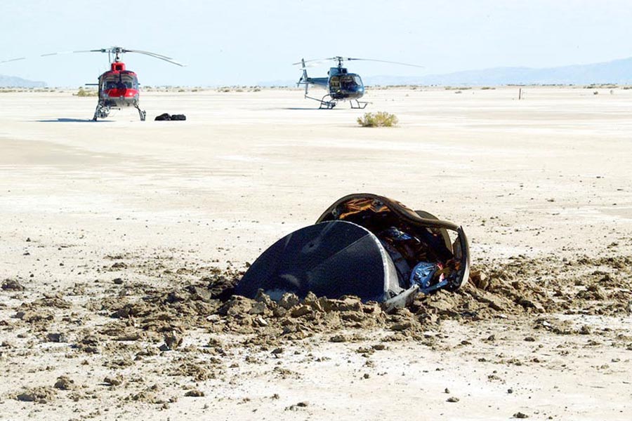 Im Vordergrund ist eine zerstörte Sonde in den Boden gerammt, im Hintergrund stehen zwei Hubschrauber auf einer sandigen Ebene.