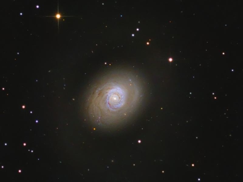 In der Mitte des Bildes mit wenigen Sternen leuchtet eine Galaxie, die wir von oben sehen, die mit ihren wenigen Spiralarmen wie eine Fläche wirkt.