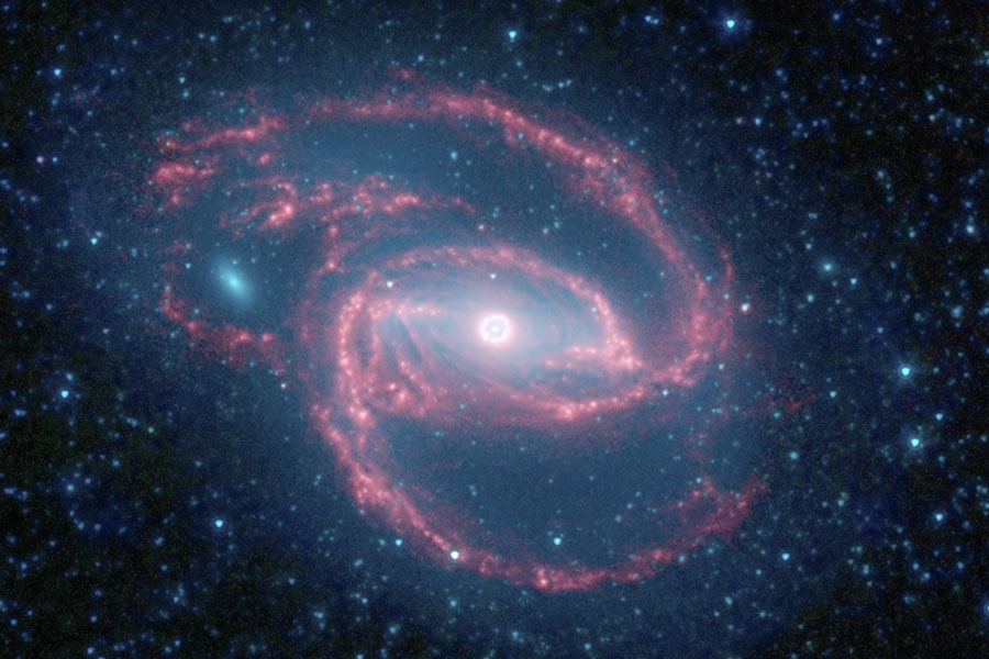 Die Galaxie im Bild hat einen sehr hellen, kleinen Kern, von einem Balken gehen rosafarbene Spiralarme aus, die lose gewunden, aber klar definiert sind.