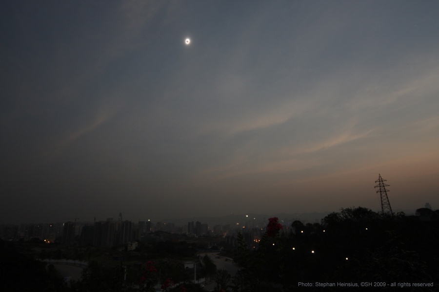 An einem dämmrigen Tageshimmel leuchtet die vom Mond verdunkelte Sonne mit einer hellen Korona außen herum.