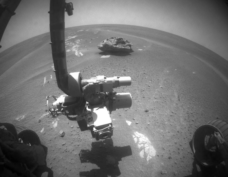 Ins Bild ragt ein Instrumentenarm eines Marsrovers, im Hintergrund ist die stark gekrümmte Oberfläche des Mars zu sehen.