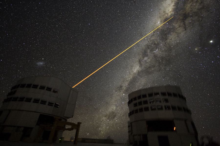 Zwischen zwei Teleskoptürmen steigt die Milchstraße mit markanten Staubwolken auf, vom linken Teleskop leuchtet ein Laserstrahl in die Sternwolken.