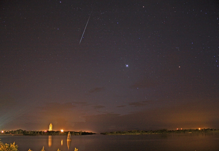 Nächtlicher Blick über ein Gewässer, am hinteren Ufer startet eine Raumfähre, von oben fällt ein Meteor vom dunklen Himmel.