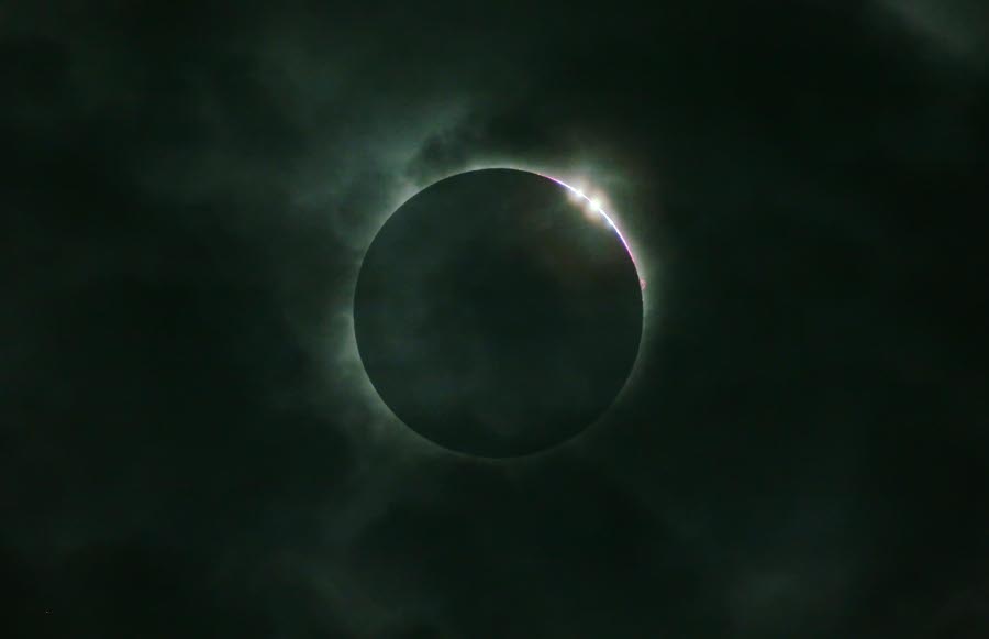 Um die dunkle kreisförmige Silhouette des Mondes leuchtet ein zarter Schimmer, rechts oben leuchten zwei helle Bailysche Perlen.