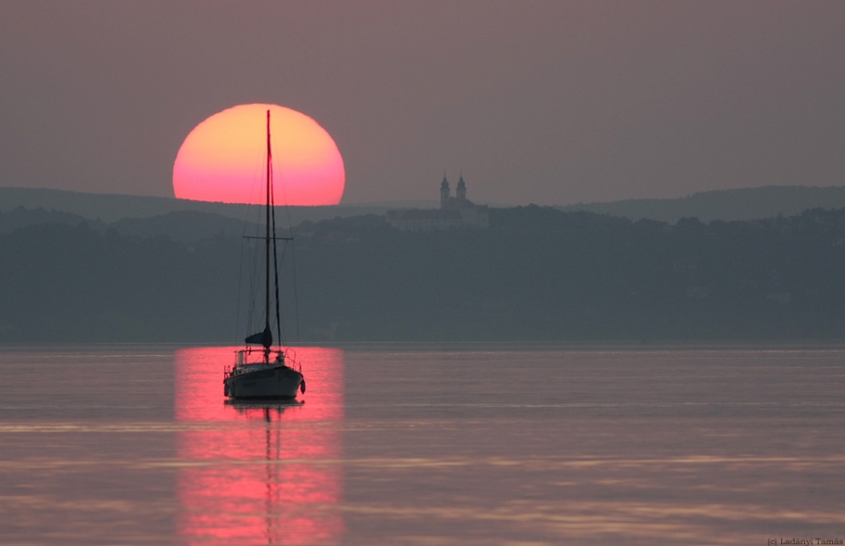 Die untergehende, orangerote Sonne spiegelt sich im Wasser, auf dem ein Boot an derselben Stelle schwimmt.
