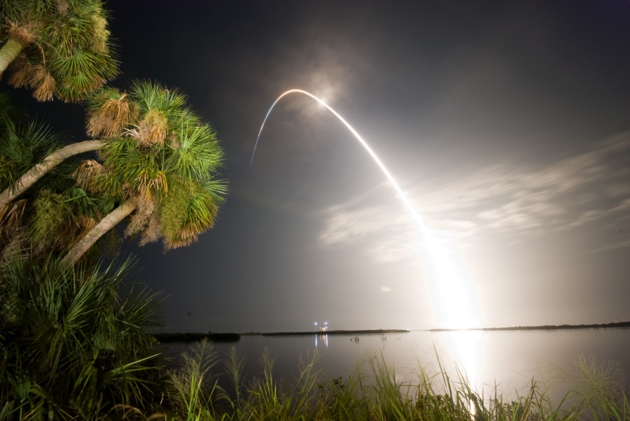 Hinter einem Gewässer mit Palmen am Ufer leuchtet die Spur eines Raketenstarts, die sich im Wasser spiegelt. Am hinteren Ufer ist der Startrampenkomplex am Kennedy Space Center zu sehen.