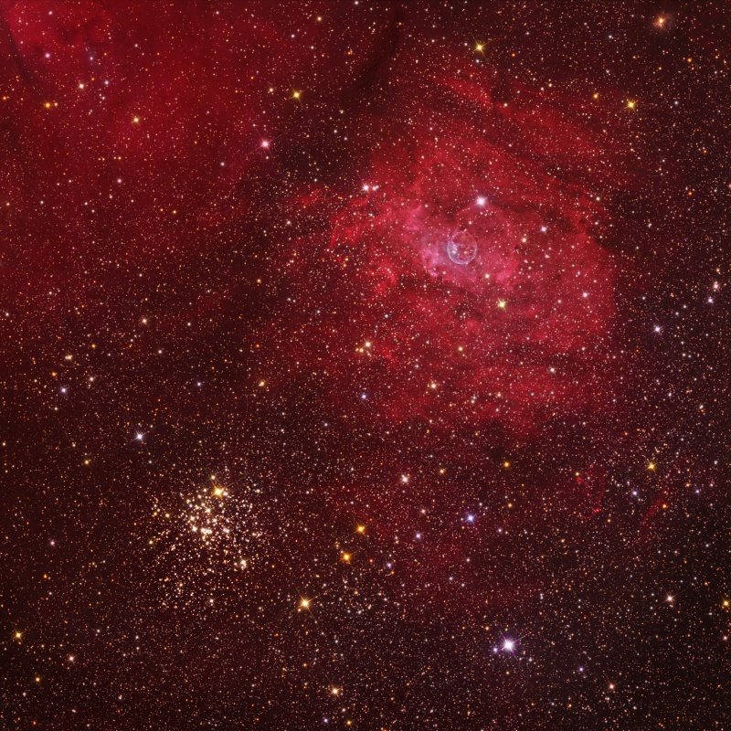 Das sternenriche Bild ist von Nebeln durchzogen. Links unten befindet sich ein Sternhaufen, Über der Mitte leuchtet ein rötlicher Nebel, in dessen Mitte sich ein kleiner blasenförmiger Nebel befindet.