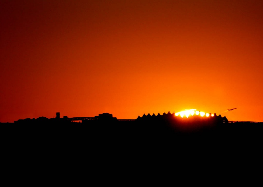 Ein kleiner Teil der Sonne ragt über den Horizont, der Himmel ist orangefarben, am Horizont ragt die Silhouette des Flughafens Denver auf.