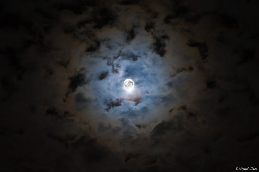 Der Mond in der Mitte ist von Wolken umgeben, die nur zu sehen sind, wo er sie beleuchtet: In der Mitte bläulich, auslaufend nach braun. Darum herum ist das Bild dunkel.