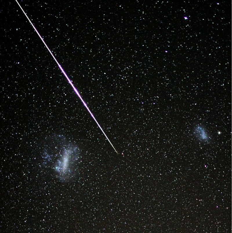 Am sterngesprenkelten Himmel zischt von links oben diagonal ein Meteor durchs Bild. Links unten leuchtet die große Magellansche Wolke, rechts auf halber Höhe die kleine Magellansche Wolke, zwei graue Nebelfleckchen.