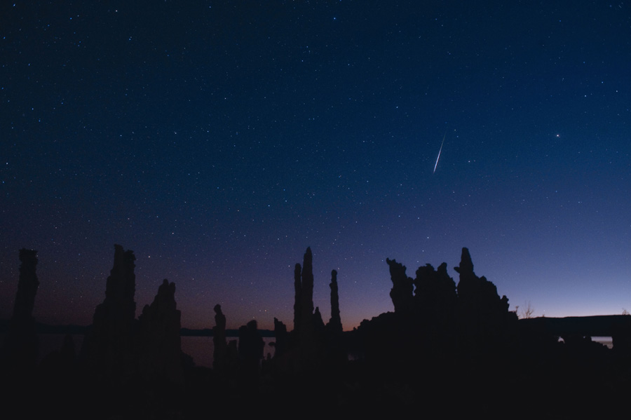 Hinter einer dunklen Silhouette des Horizonts mit Bäumen ist der Himmel sehr dunkelblau. Rechts auf neben der Mitte leuchtet die Spur eines Meteors.