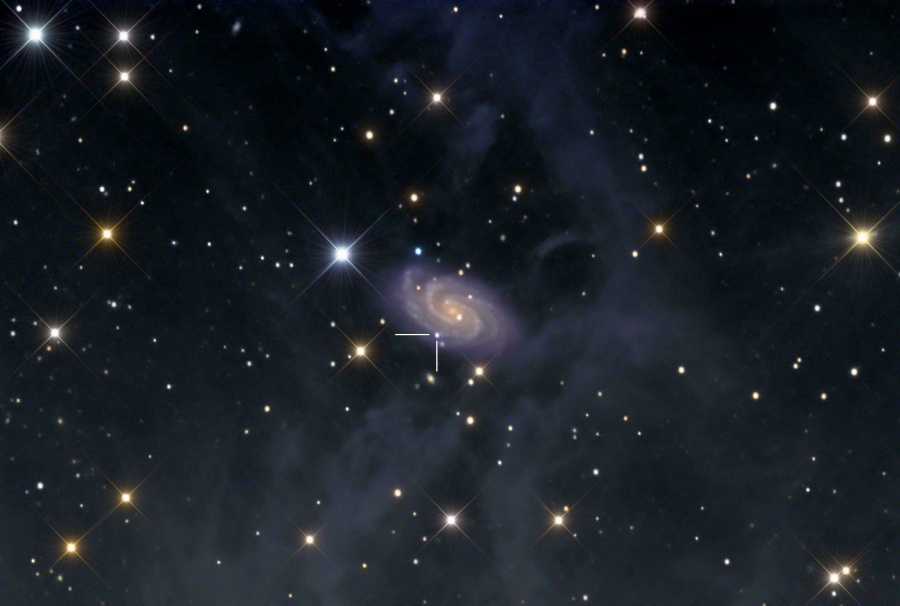 Im Bild sind wenige, aber markante Sterne verteilt, eine davon mit Zacken. In der Mitte leuchtet eine kleine Spiralgalaxie zwischen zarten Nebeln, links unten ist ein heller Lichtpunkt markiert - eine Supernova.