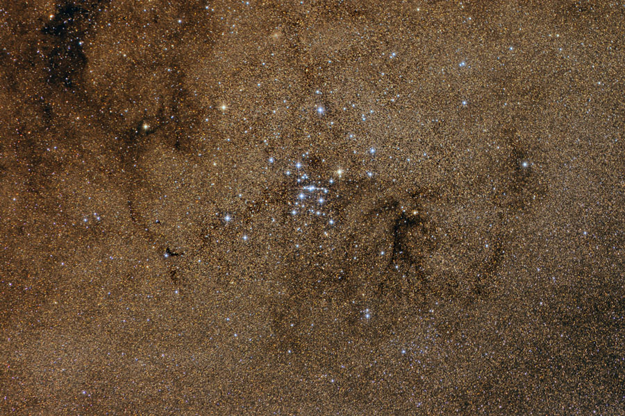 Das Bild in braunen Farbtönen ist dicht mit Sternen besetzt, dazwischen einige dunkle Fasern, in der Mitte ist ein bläulicher Sternhaufen.