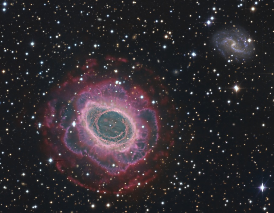 Links unten ist ein großer, runder Nebel, der an eine Rosette erinnert. Rechts oben leuchtet hinter einem Vordergrund mit hellen Sternen eine S-förmige Balkenspiralgalaxie, sie ist kleiner als der Ringnebel.