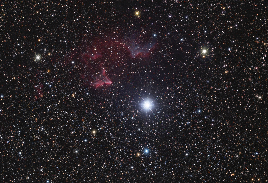 Mitten im sternbedeckten Bild leuchtet ein helles Licht, links darüber sind rötliche Nebel erkennbar.