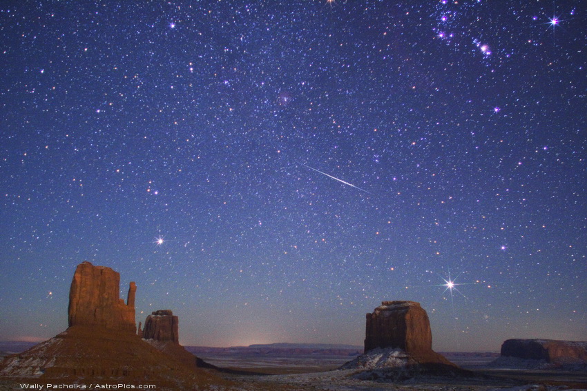 Hinter Tafelbergen im Monument Valley ist ein prächtiger Sternenhimmel mit dem Orionnebel rechts oben und einem Meteor in der Mitte.