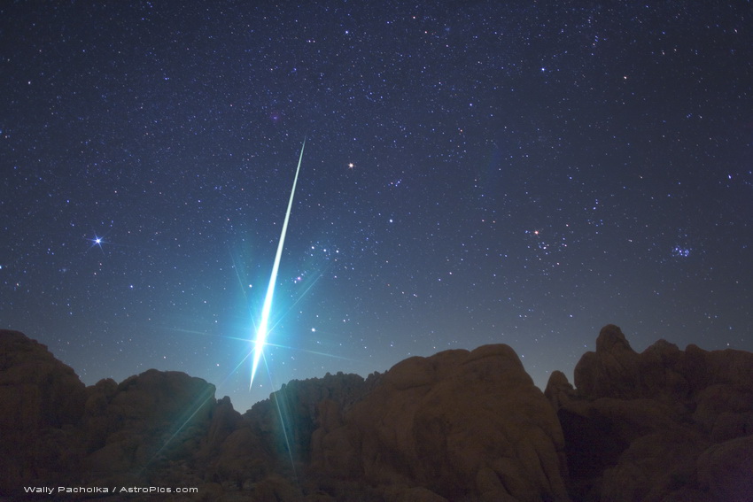 Hinter einer Silhouette mit zerklüfteten Bergen blitzt eine helle Feuerkugel vor dem Sternbild Orion auf. Rechts neben Orion sind die Hyaden und die Plejaden, links davon der helle Stern Sirius.
