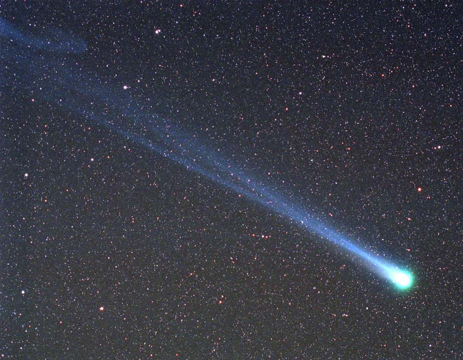 Rechts unten leuchtet der Kopf eines Kometen mit grünlichem Rand, sein Schweif reicht nach links oben über ein sterngesprenkeltes Bildfeld.
