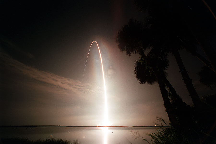 Über einem ruhigen Wasser steigt die feurige Parabel der Abgase eines Raketenstarts auf, rechts steht die Silhuoette von Palmen.