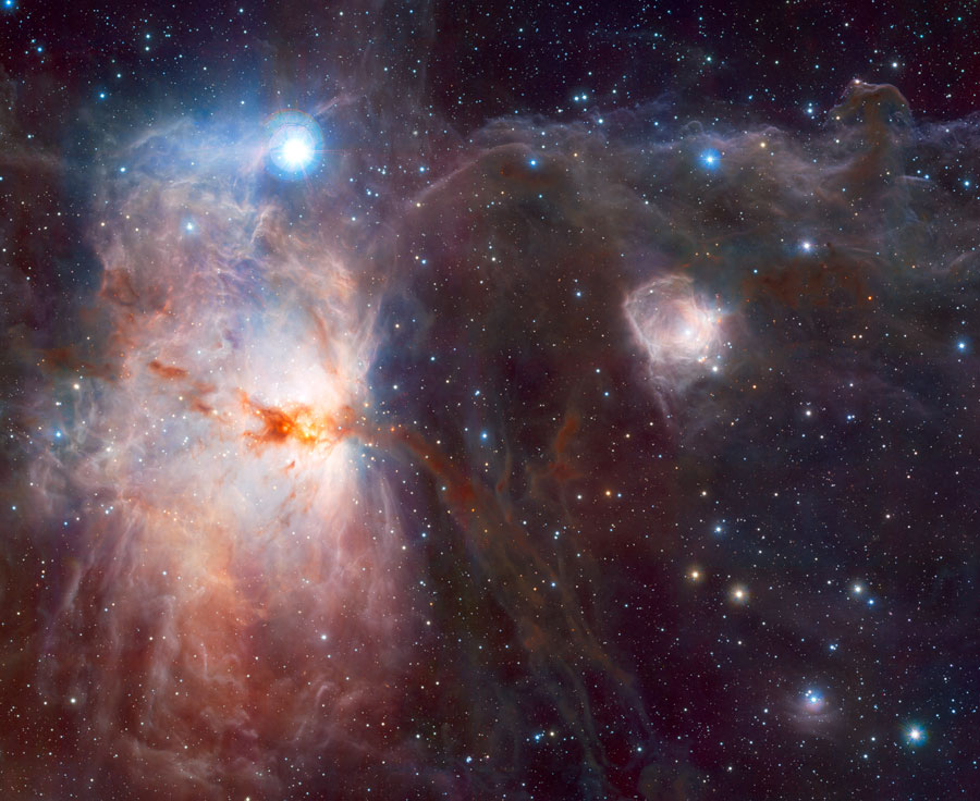 Links im Bild leuchtet der Flammennebel, oberhalb ist ein blau leuchtender Stern. Rechts sind einige Staubwolken erkennbar, eine kleine leuchtet ebenfalls. Auch Sterne sind im Hintergrund und im Vordergrund verteilt.