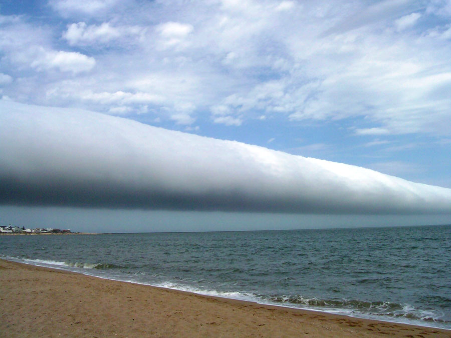 Links ist ein Sandstrand, von dem aus nach rechts ein unruhiges Meer bis zum Horizont reicht. Über dem Horizont reicht eine walzenförmige Wolke quer durch das ganze Bild.