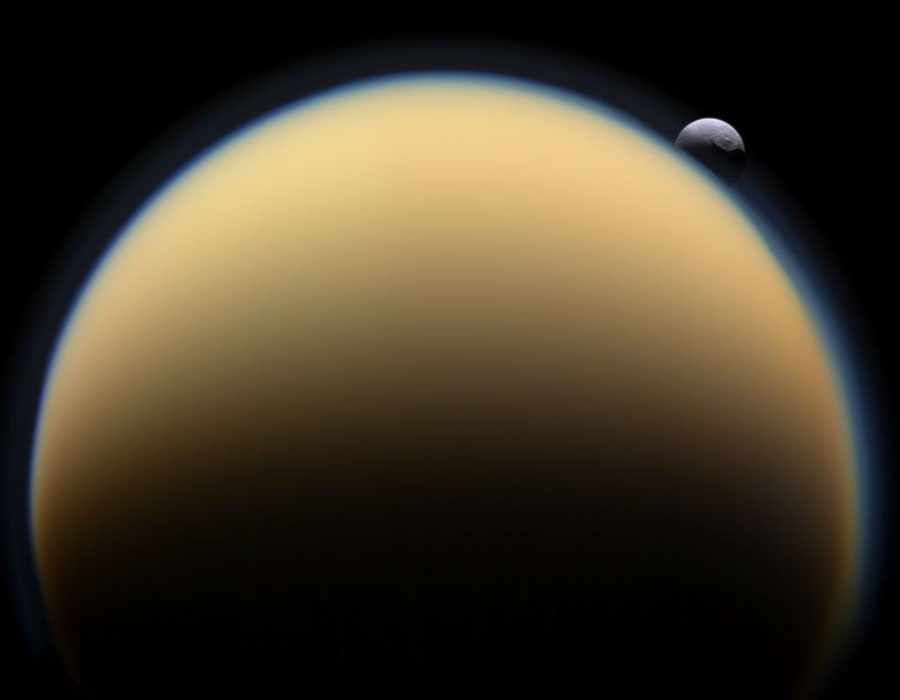 Im Vordergrund ragt die Sichel des Saturnmondes Titan nach oben hoch, sie ist orange getönt mit einem bläulichen Rand. Rechts darüber ragt der Mond Tethys über den Rand.