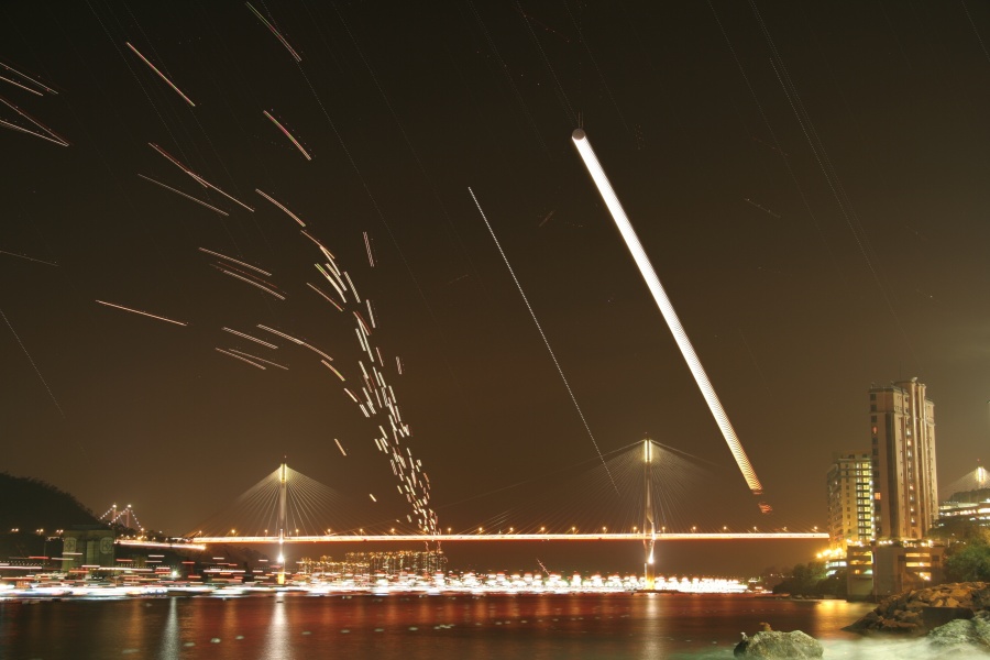 Über einer hell beleuchteten Stadt blicken wir über ein Gewässer auf eine Brücke. Links steigen Lichtstreifen auf, rechts verläuft ein breiter heller Streifen schräg nach links zur oberen Mitte. Ganz rechts stehen Hochhäuser.